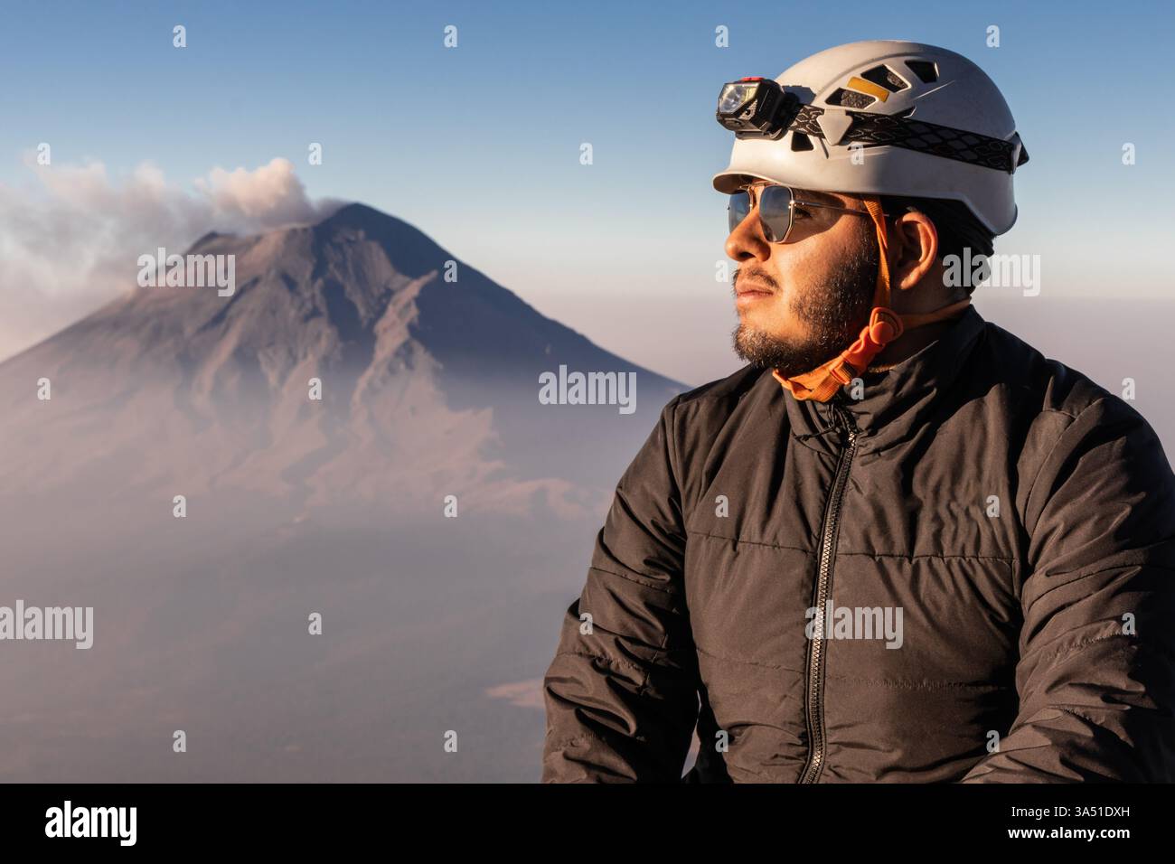 Ein männlicher Wanderer mit Helm und Sonnenbrille bewundert den Blick auf einen rauchenden Vulkan bei Sonnenaufgang an den Hängen des Iztaccihuatl. Dieses dramatische Reisebild erfasst Abenteuer, raue Berglandschaften und Outdoor-Entdeckungen für Natur- und Reisekampagnen. Stockfoto