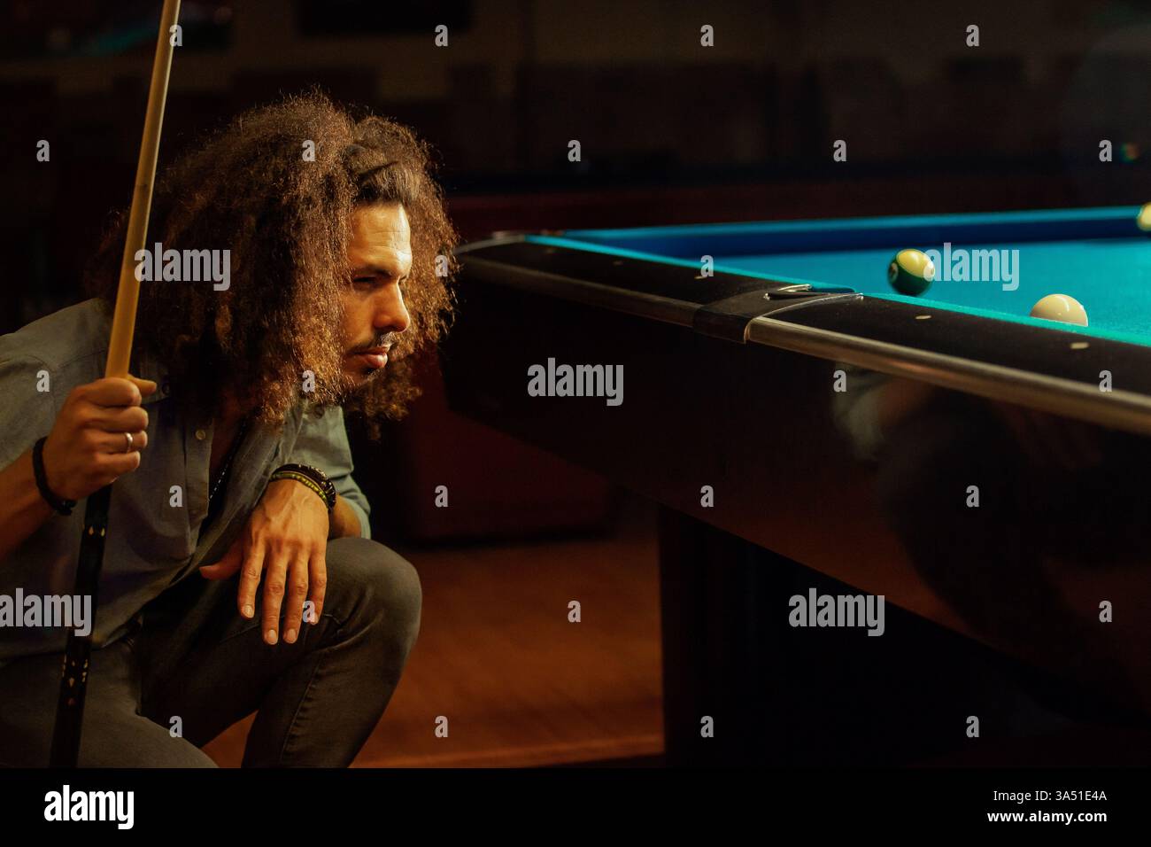 Konzentrierter hispanischer Mann hockt sich, um einen Schuss in einer Billardhalle zu sehen und konzentriert sich auf das Billardspiel. Dieser dynamische Sportereignis eignet sich für Freizeit, Unterhaltungsangebote und Billardaktionen. Stockfoto