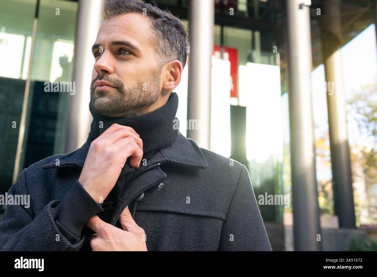 Porträt eines spanischsprachigen Geschäftsmannes, der seinen Mantel auf einer sonnigen Stadtstraße anpasst. Die Tageslichtaufnahme für den Außenbereich vermittelt Professionalität und Konzentration. Ideal für Unternehmenskampagnen, Stadtleben und Branding. Stockfoto