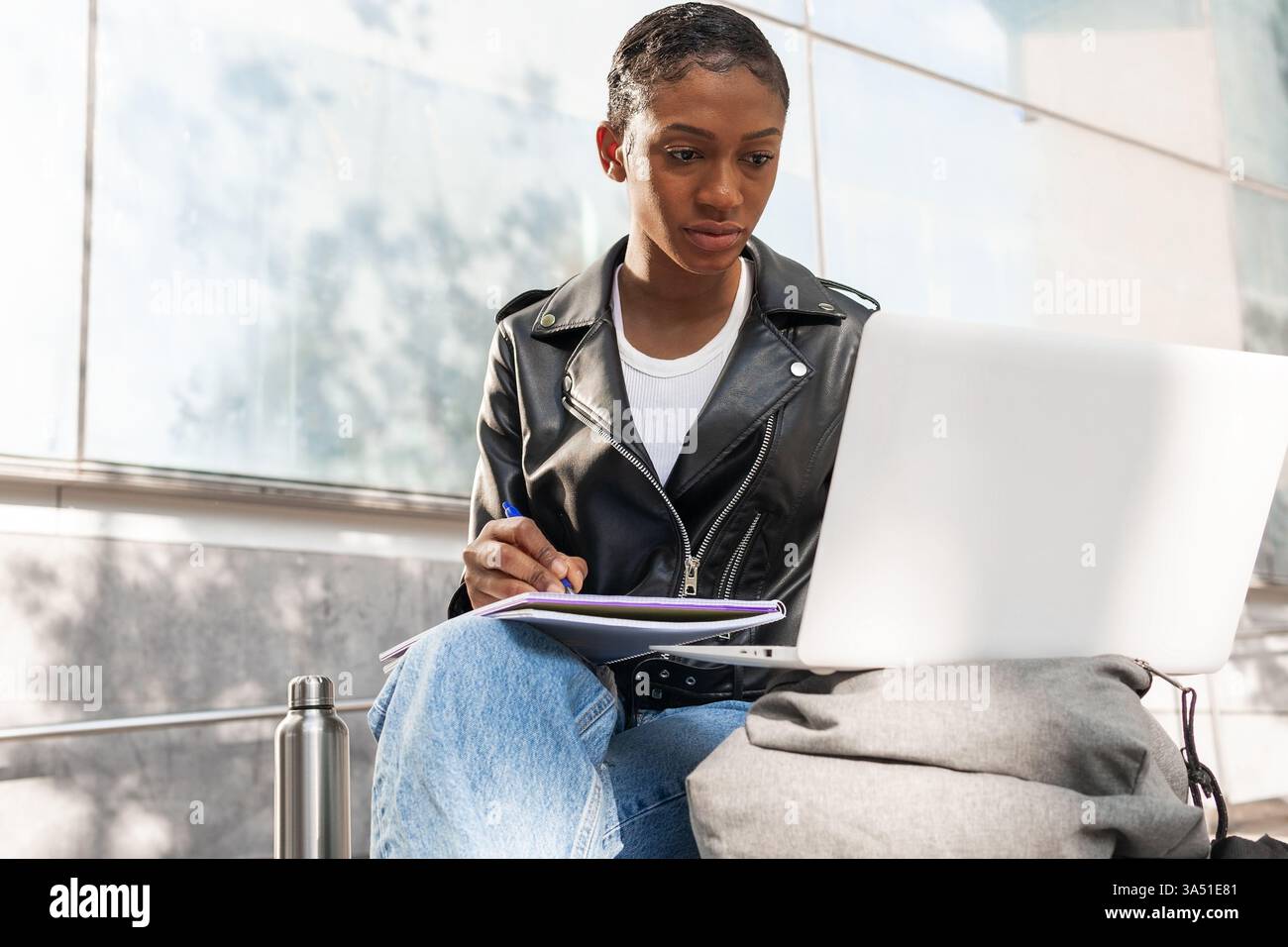 Schwarze Frau schreibt in einem Notizbuch, während sie draußen einen Laptop ansieht. Das Bild kombiniert die Produktivität von Studien und Außenanwendungen in einer städtischen Umgebung. Nützlich für das Lernen im Freien, die Arbeit an der Ferne und die Darstellung moderner Lifestyle. Stockfoto