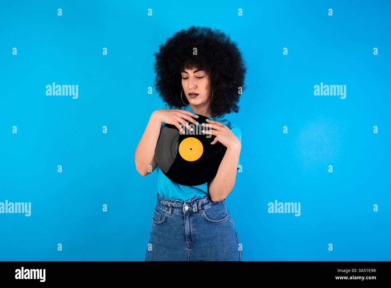 Ernsthafte Hispanic Woman With an Afro umarmt eine Retro-Schallplatte und blickt vor blauem Hintergrund nach unten. Ideal für Musik, Vintage-Stil und nostalgisches Branding. Stockfoto