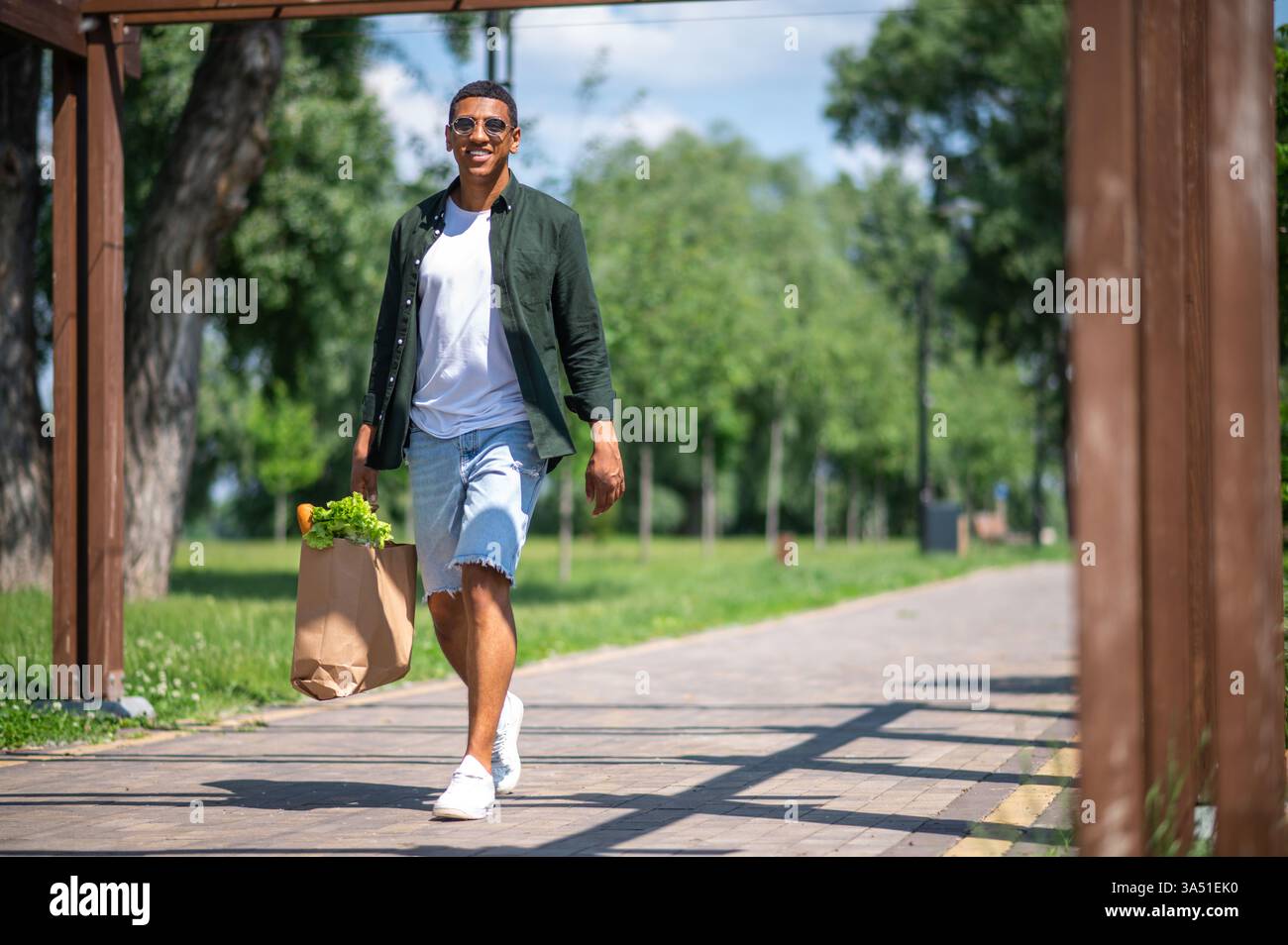 Ein lächelnder Afroamerikaner mit Sonnenbrille und einer Tasche Essen, während er durch einen sonnigen Park schlendert. Ein fröhliches Outdoor-Lifestyle-Bild mit natürlichem Licht, Grün und entspannter Stimmung, ideal für Wellness-, Food- oder Freizeitkampagnen. Geeignet für redaktionelle oder kommerzielle Anwendungen mit positiven Vibes und Outdoor-Aktivitäten. Stockfoto