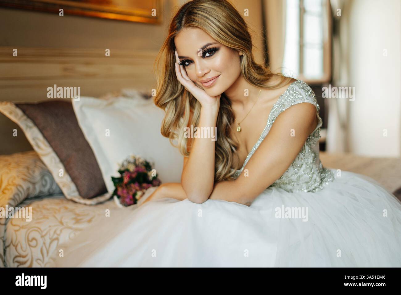 Fröhliche Braut in einem eleganten weißen Hochzeitskleid sitzt auf einem Bett mit einem Blumenstrauß in einem sanft beleuchteten Schlafzimmer. Dieses Brautporträt fängt Romantik und Feier mit intimer Inneneinrichtung ein. Perfekt für Hochzeitskampagnen, Brautmode und Hochzeitsplanungsfunktionen. Stockfoto