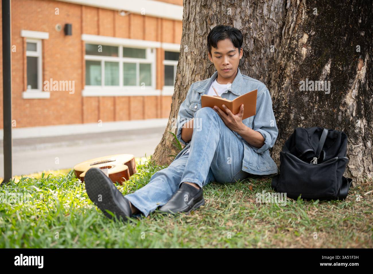 Konzentrierter asiatischer Student liest ein Buch unter einem Baum in einem Campus-Park. Lässige Outdoor-Lernszene für Schülerleben, Bildung und Lernthemen. Ideal für Universitäten, Campus und Inspiration. Stockfoto