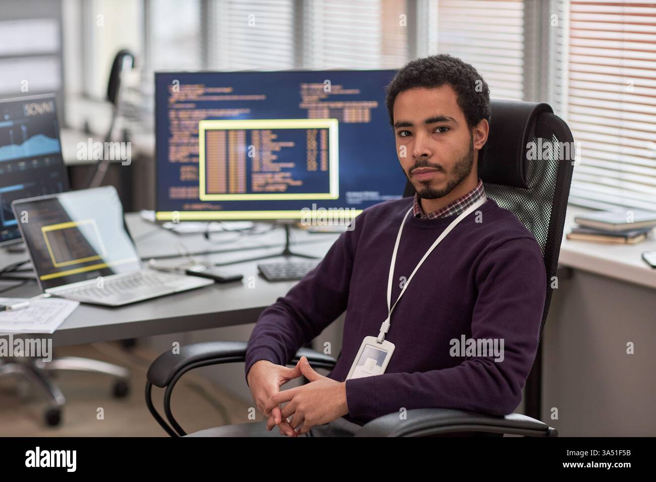 Porträt eines männlichen Datenwissenschaftlers, der in die Kamera schaut, während er im IT-Entwicklungsbüro mit Computern im Hintergrund am Schreibtisch sitzt Stockfoto