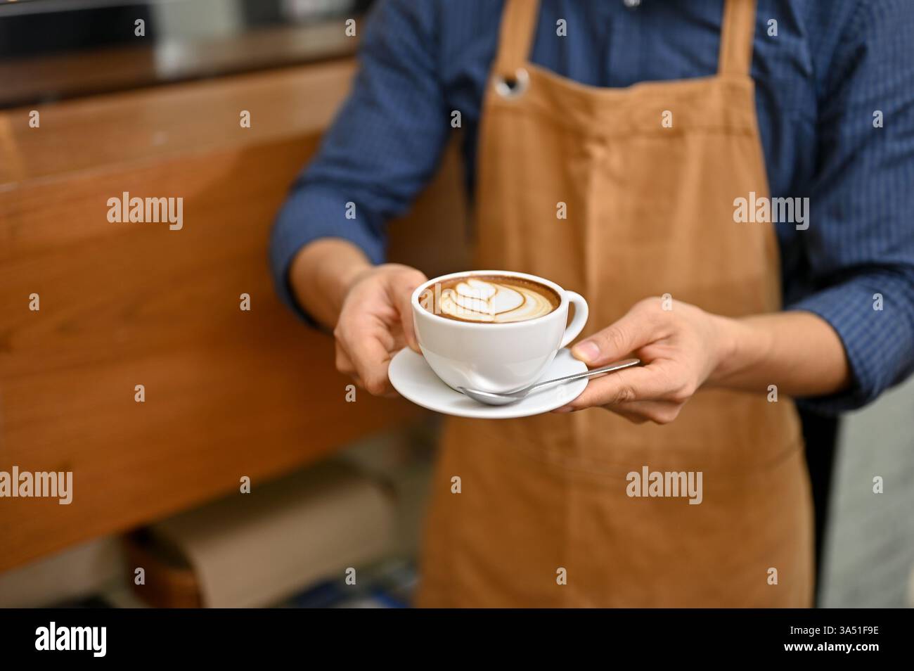 Männlicher Barista in einem Café, der eine Tasse mit Latte Art vor der Theke präsentiert. Die gekürzte Komposition betont Latte Art, Café-Atmosphäre und fachkundigen Service. Ideal für Lebensmittel- und Getränkemarketing. Stockfoto