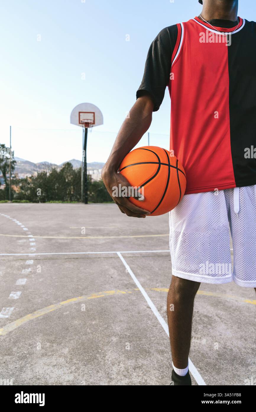 Schwarzer männlicher Athlet in Uniform steht mit einem Basketball auf einem Platz. Dynamisch und bereit für Action, passt dieses Bild zu Sport-, Trainings- und sportlichen Lifestyle-Themen. Stockfoto