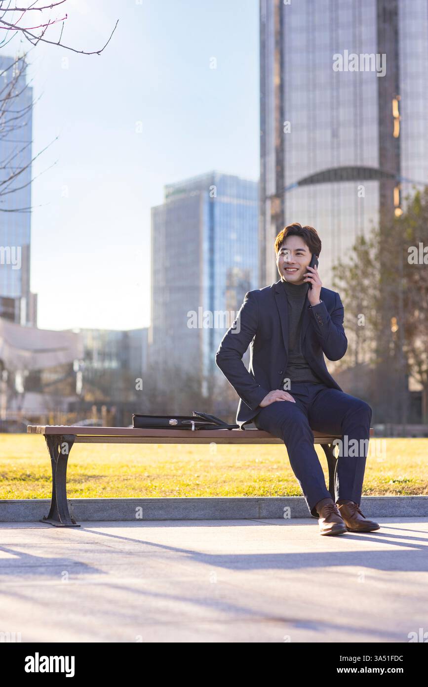 Fröhlicher chinesischer Geschäftsmann, der auf seinem Handy spricht, im Freien mit Stadtkulisse festgehalten. Perfekt für Unternehmens-, Technologie- oder Geschäftskommunikationsthemen. Stockfoto