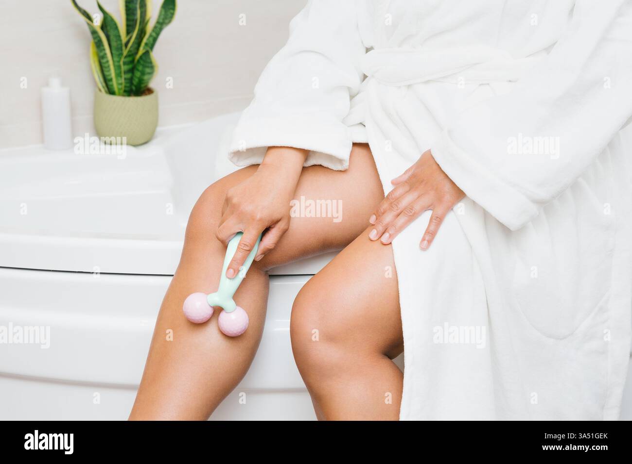 Hispanische Frau im Bademantel, die Beine mit Roller massiert, während sie zu Hause auf der Badewanne sitzt Stockfoto