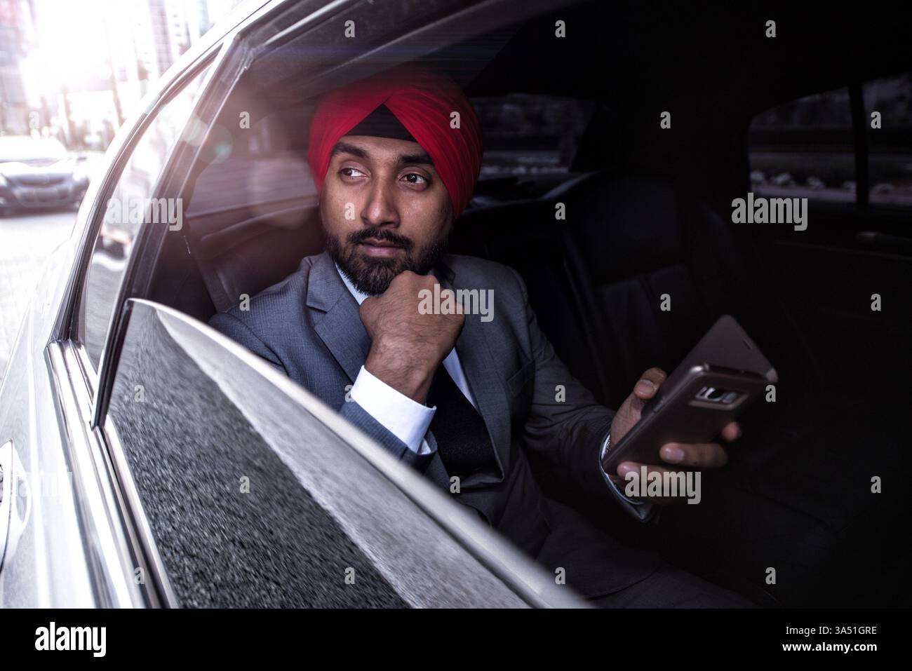 Ein indischer Geschäftsmann mit Turban sitzt während der Reise in einem Auto und berührt sein Kinn in einer nachdenklichen Pose. Die Szene vermittelt geschäftliches Pendeln und professionellen Stil unterwegs. Stockfoto