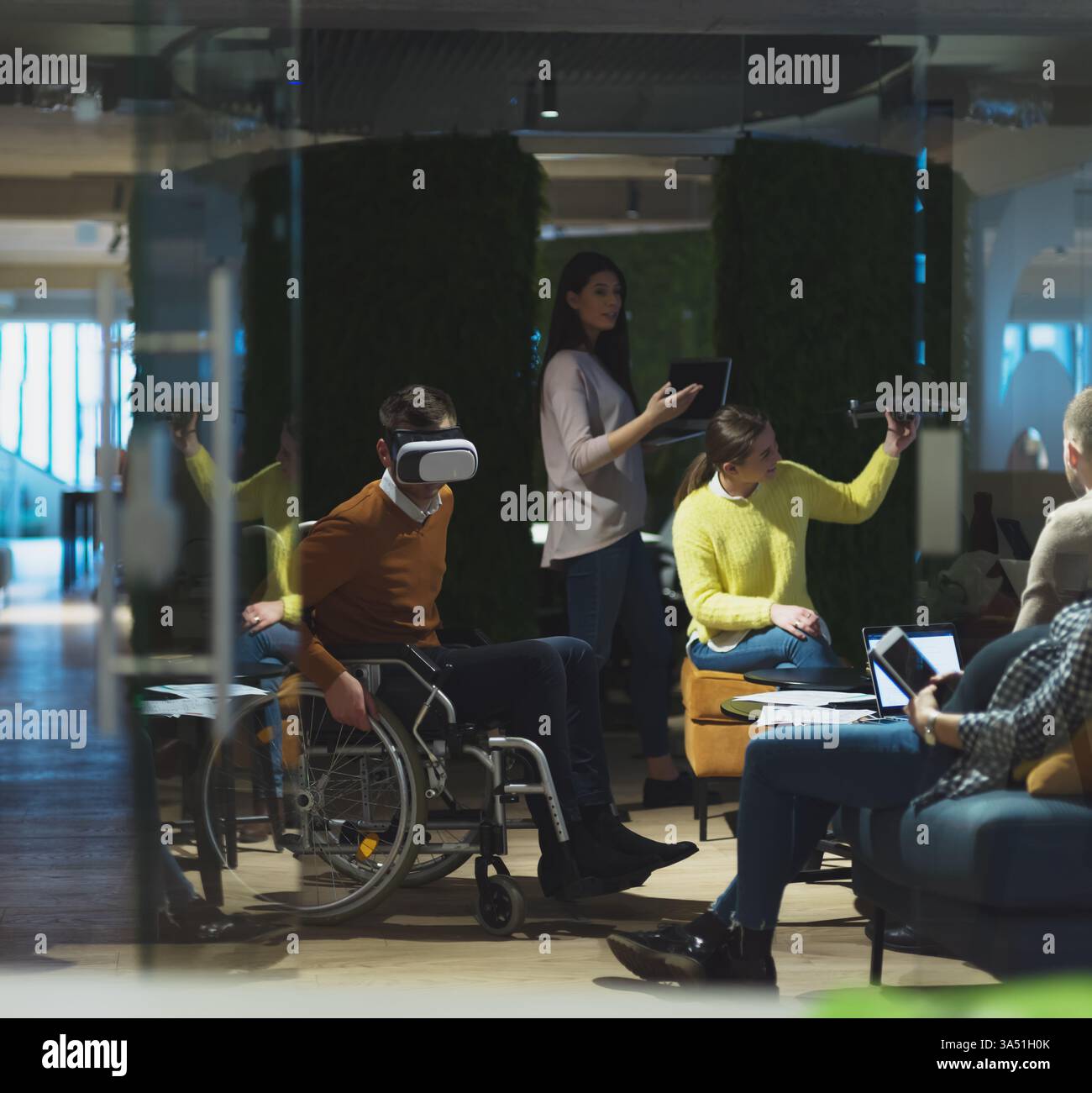 In einem modernen, offenen Coworking-Raum führt ein behinderter Geschäftsmann im Rollstuhl ein Teammeeting mit einer Virtual-Reality-Schutzbrille. Drohnenunterstützung und eine vielfältige Startup-Umgebung heben Innovation, Zusammenarbeit und integrative Arbeitsbedingungen hervor. Stockfoto