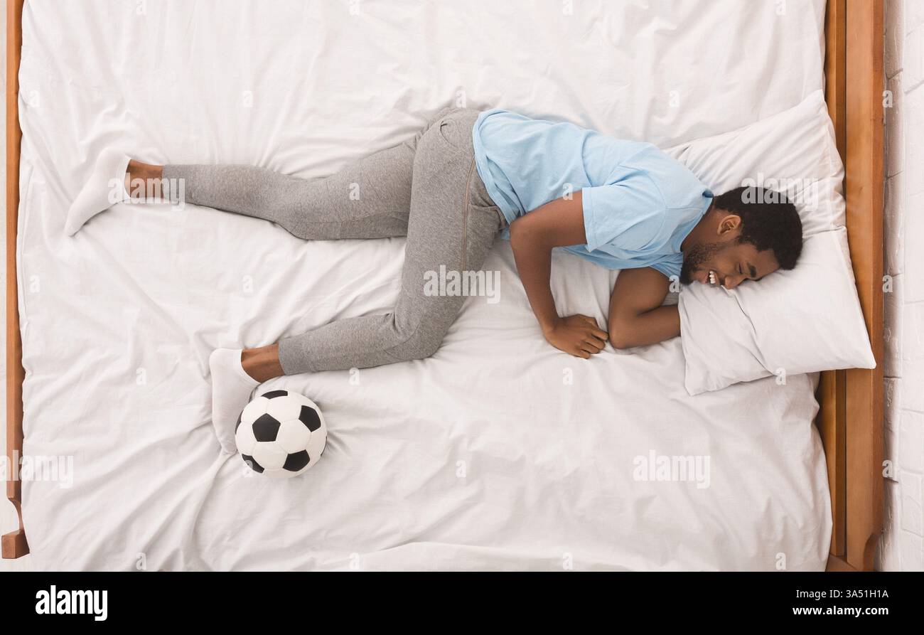 Bild von oben von einem lachenden Schwarzen, der mit einem Fußball schläft und davon träumt, Fußball zu spielen. Dieses verspielte Bild eignet sich für Sport, Fankultur und Ruhemomente in der Sportfotografie. Ideal für Ideen zu Entspannung, Träumen und sportlichem Lifestyle. Stockfoto