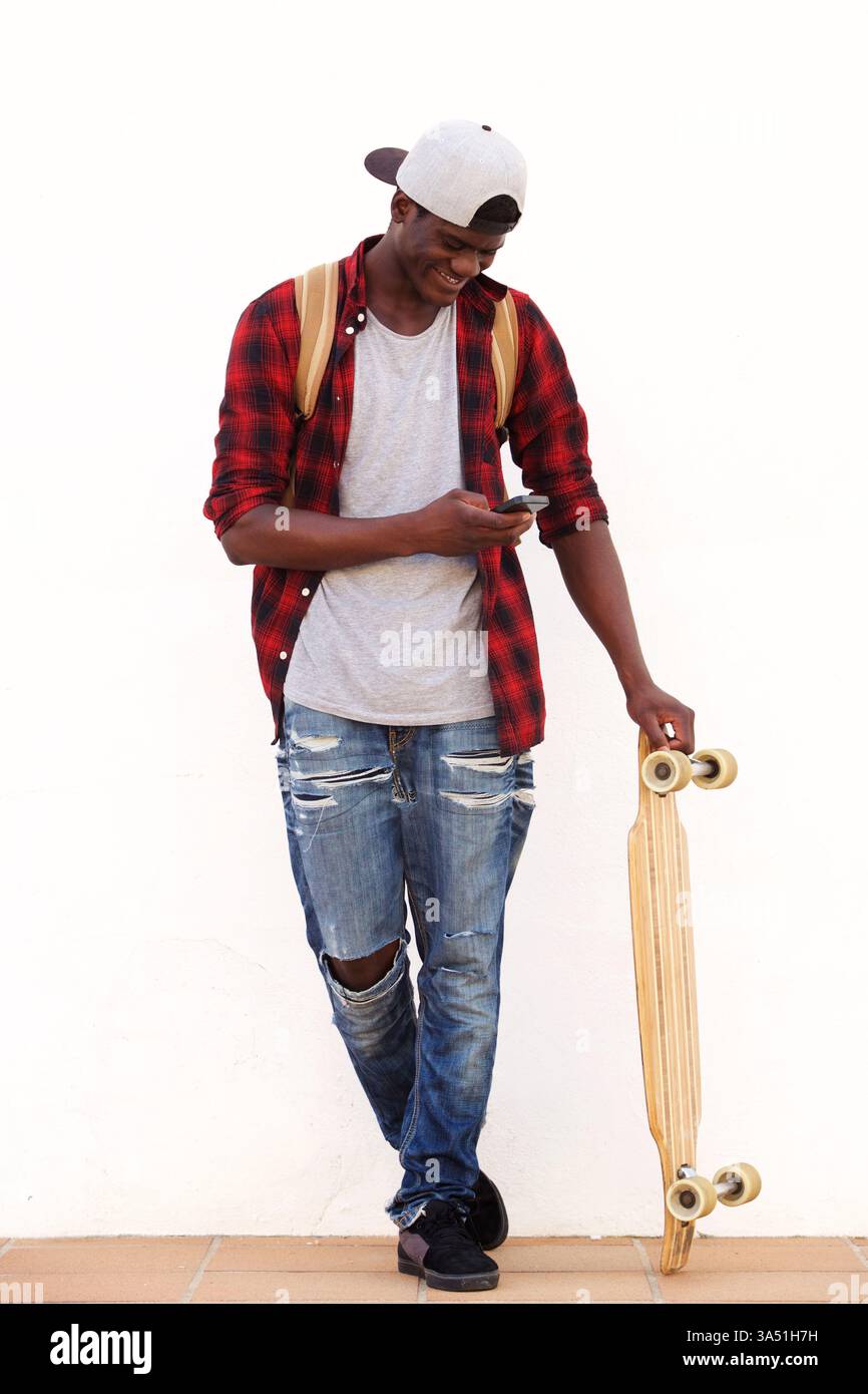 Ganzkörperporträt eines glücklichen jungen Schwarzen Mannes mit einem Skateboard, der sein Handy ansieht und lächelt. Der lässige, moderne Stil vermittelt Jugend, Mode und einen aktiven Lebensstil. Geeignet für Street-Style, Teenager-Lifestyle und Stadtfotografie. Stockfoto