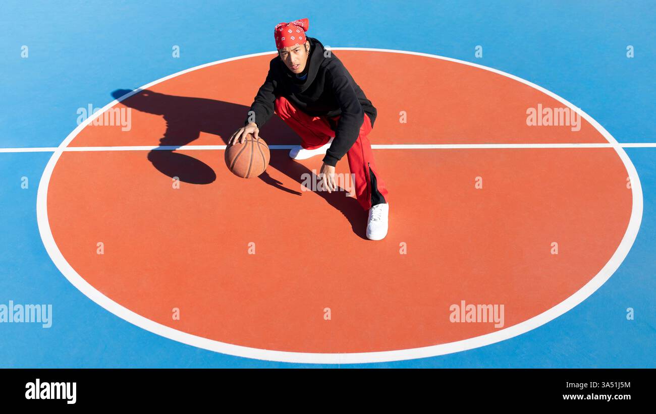 Asiatischer Mann mit rotem Bandana Dribbling Ball während er sich auf dem Sportplatz an sonnigen Tagen hockt Stockfoto