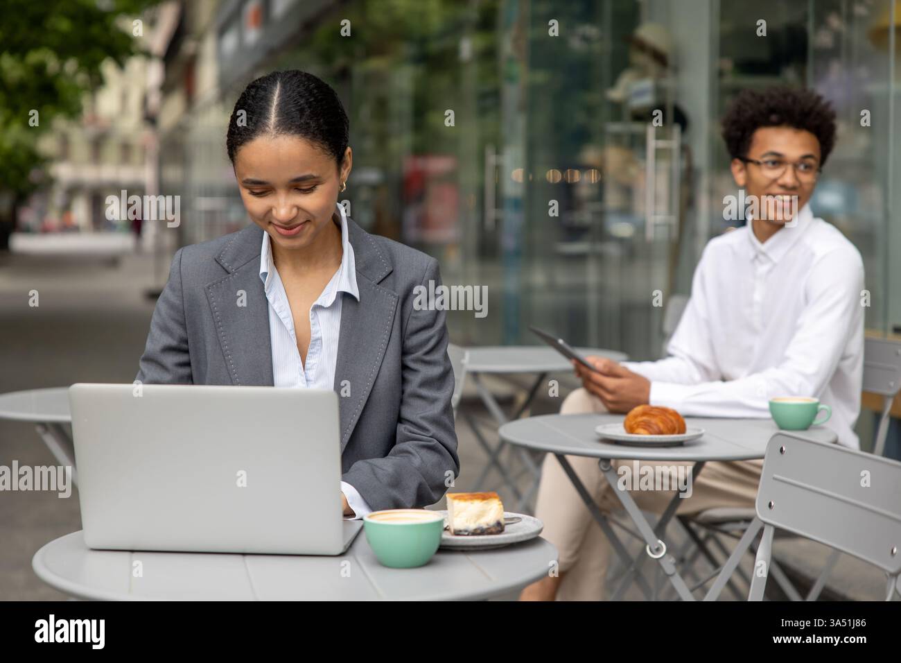 Schwarze Geschäftsfrau, die tagsüber einen Laptop in einem Café im Freien bei einer Tasse Kaffee benutzt, mit einem männlichen Kollegen in der Nähe. Steht für moderne flexible Arbeit, Remote Collaboration und tagtägliche Geschäftsgespräche. Stockfoto