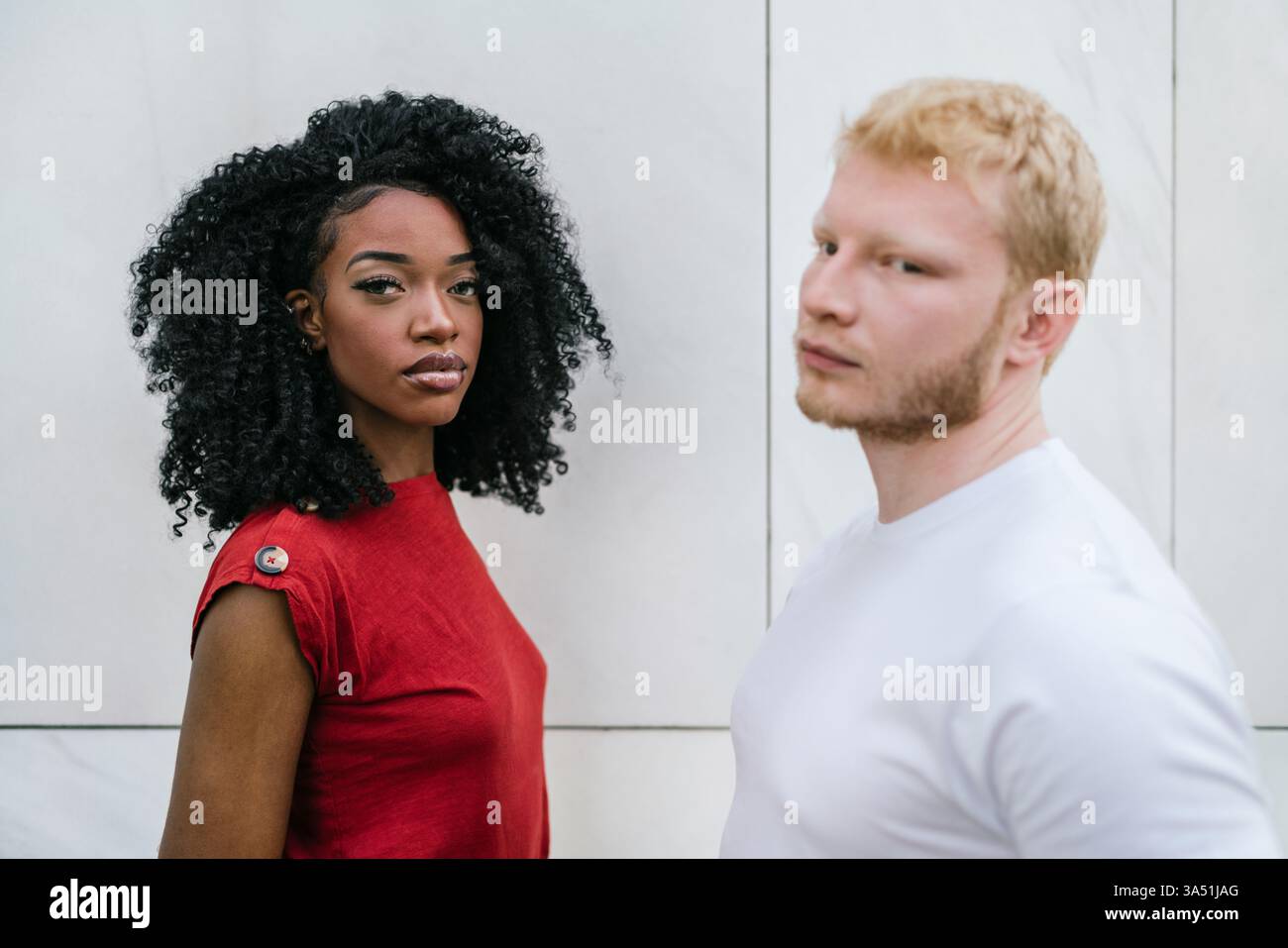 Seitenansicht eines vielseitigen Paares im legeren Stil. Die afroamerikanische Freundin mit Make-up schaut in die Kamera, während ihr bärtiger Freund in einem weißen T-Shirt in nachdenklicher Stimmung in der Nähe steht. Dieses Bild zeigt eine zeitgemäße Beziehungsdynamik, die für Lifestyle und Paare geeignet ist. Stockfoto