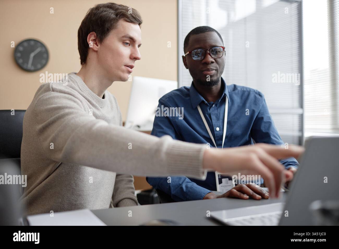 Taille-up-Aufnahme eines jungen kaukasischen Mannes, der auf einen Laptop-Bildschirm zeigt, während er mit einem afroamerikanischen Kollegen in einem modernen Büro sprach. Eine vielfältige, kollaborative Arbeitsplatzszene, die sich für Technologie-, IT- und Teamwork-Kampagnen eignet. Stockfoto