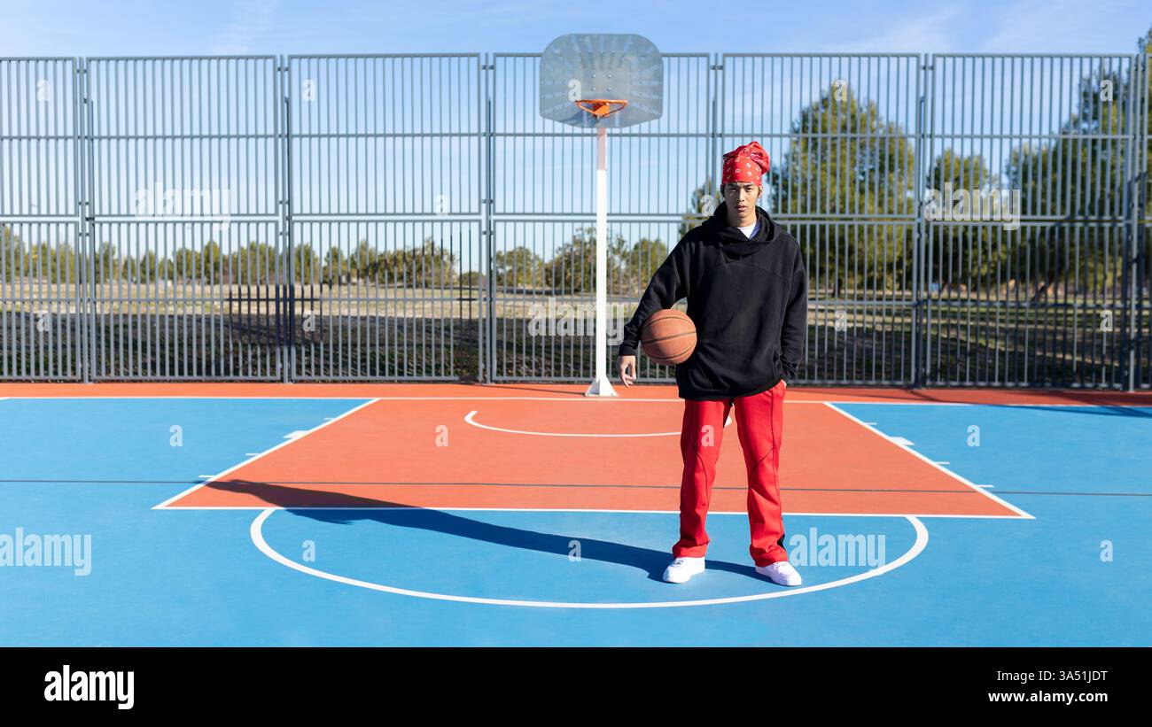 Ganzkörperaufnahme eines ernsthaften Asiaten in Sportbekleidung, der einen Basketball auf einem Sportplatz hält. Er schaut in die Kamera, bereit für Training und Wettkampf. Ein dynamisches Sportimage, das perfekt für Fitness-, Basketball- und sportliche Lifestyle-Kampagnen geeignet ist. Stockfoto