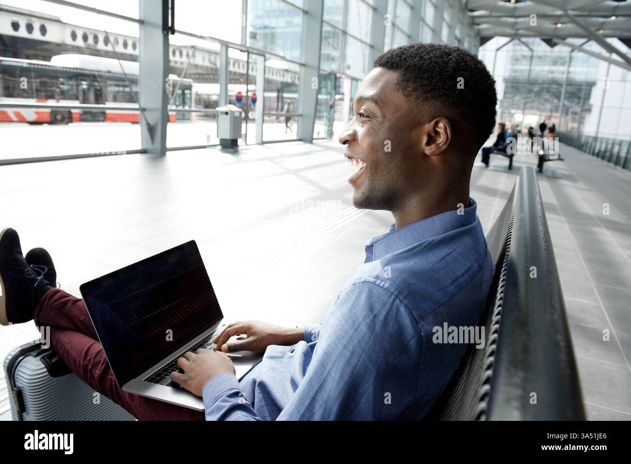 Afroamerikanischer Geschäftsmann wartet auf einer Bank mit Taschen und Laptop, bereit für Reisen oder einen anstrengenden Arbeitstag. Dieser urbane Moment für unterwegs fängt einen selbstbewussten, professionellen Reisenden ein. Nützlich für Geschäfts-, Reise- und Lifestyle-Bilder. Stockfoto