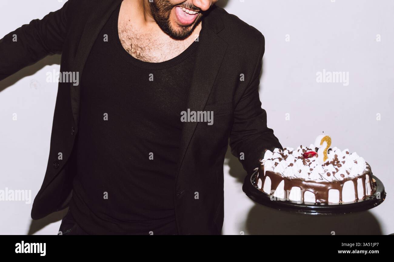 Ein lächelnder hispanischer Mann tanzt mit einer Geburtstagskuche vor einer weißen Wand und feiert einen fröhlichen Geburtstagsmoment. Diese lebhafte Szene weckt Partystimmung, Feiern und fröhliche Stimmung für Veranstaltungen und Lifestyle-Kampagnen. Stockfoto