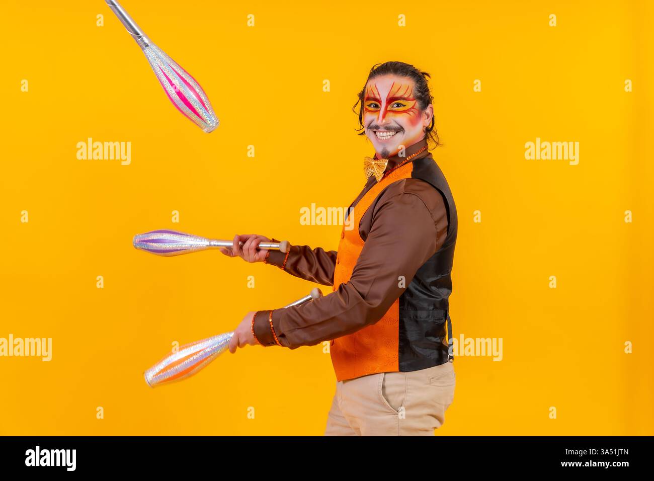 Lebendige Studioaufnahme eines Jongleurs in einer Weste und Make-up-Jonglierer vor einem kräftigen gelben Hintergrund. Das Bild vermittelt Energie, Leistung und spielerische Unterhaltung, die für Zirkus-, Party- oder Unterhaltungskampagnen geeignet ist. Eine auffällige Komposition, die sich ideal für Werbematerial mit Solo-Künstlern eignet. Stockfoto