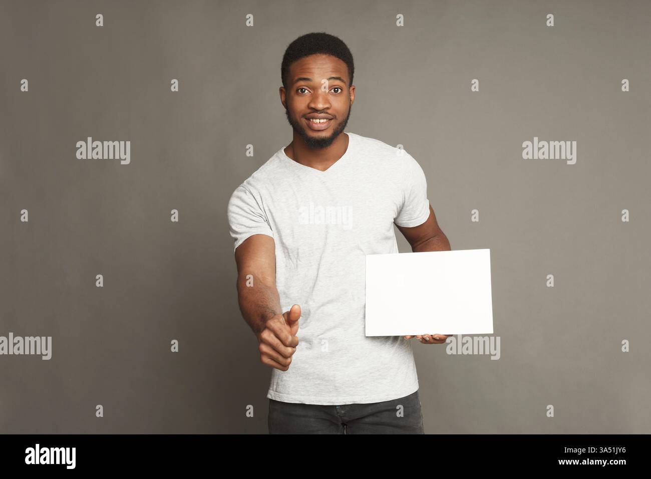 Junger afroamerikanischer Mann, der lächelt und einen Daumen hoch hält, während er eine leere Werbetafel auf grauem Hintergrund hält. Ideal für Werbung, Werbeaktionen und Layouts im Kopierbereich. Ideal für Kampagnen, die einen positiven, zugänglichen Sprecher benötigen. Stockfoto