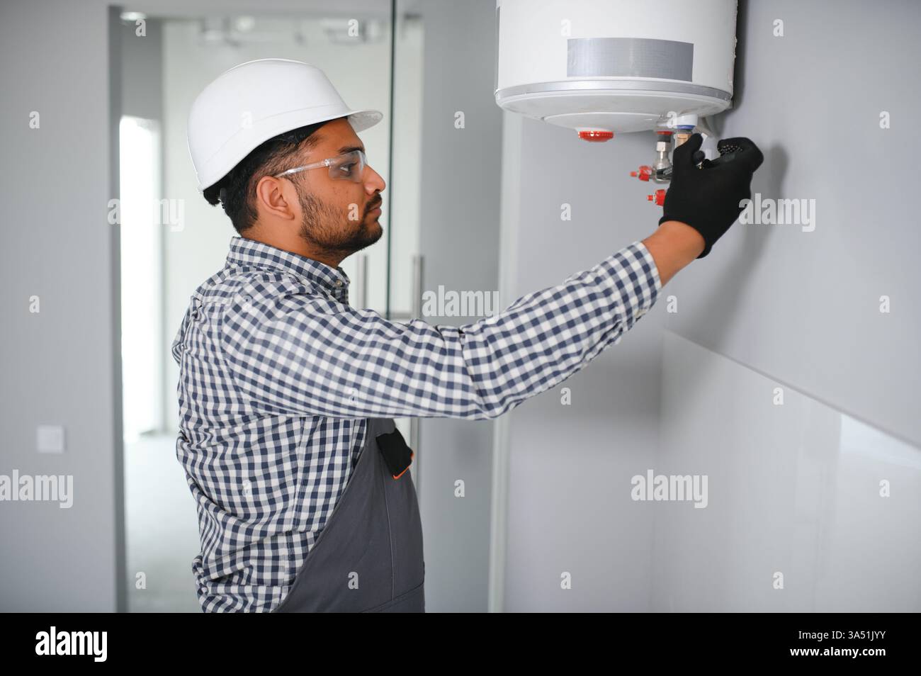 Indischer männlicher Klempner in Uniform und Schutzhelm, der an einem Heizsystem im Badezimmer arbeitet. Ein professioneller Reparaturspezialist mit Fokus auf HLK. Geeignet für Hausverbesserungen, Sanitär- und Heizungswartungskampagnen. Stockfoto