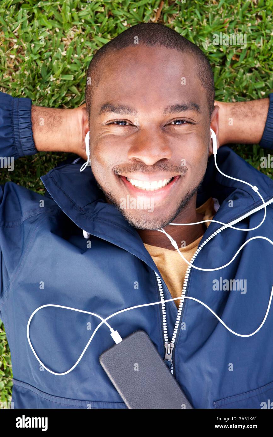 Von oben liegt ein lächelnder junger Schwarzer auf dem Gras und hört Musik mit Ohrhörern. Die Outdoor-Szene vermittelt unbeschwerte Freizeit und moderne Konnektivität. Geeignet für Lifestyle, Musik und Outdoor-Aktivitäten. Stockfoto