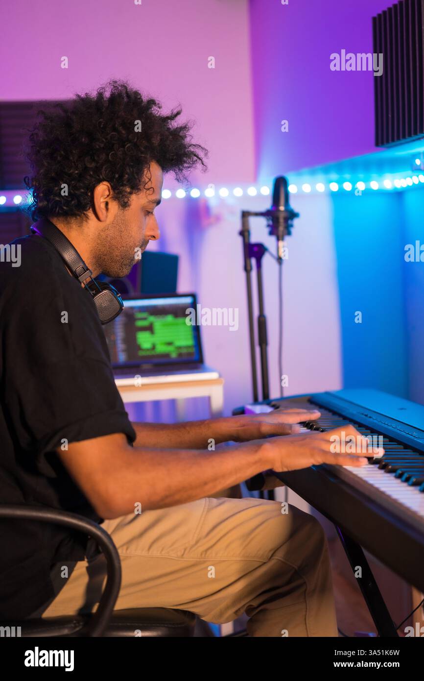 Vertikale Aufnahme eines Pianisten in einem Aufnahmestudio, der ein neues Lied aufnimmt. Neonlicht und professionelle Studioausstattung sorgen für eine kreative Musikproduktionsstimmung. Geeignet für Grafikprodukte in der Musikindustrie, Aufnahmestudios und Künstler-Workflows. Stockfoto