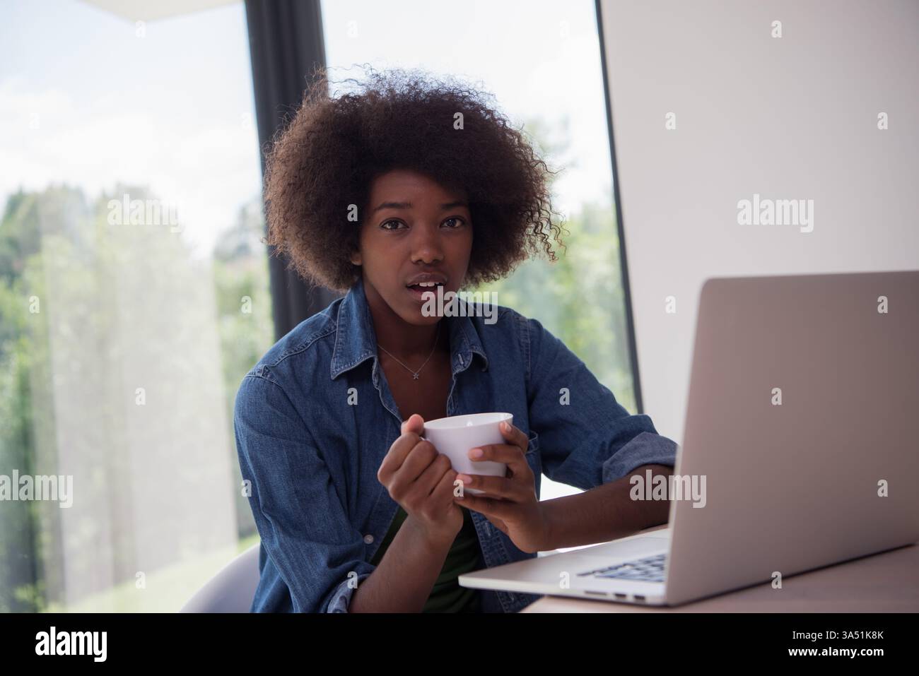 Schwarze Geschäftsfrau, die von zu Hause aus arbeitet, an einem Tisch mit Laptop und einer Tasse Kaffee sitzt. Perfekt für Fernarbeit, Home Office und digitale Unternehmensführung. Vermittelt Konzentration und berufliche Aktivität in einer komfortablen Wohnumgebung. Stockfoto