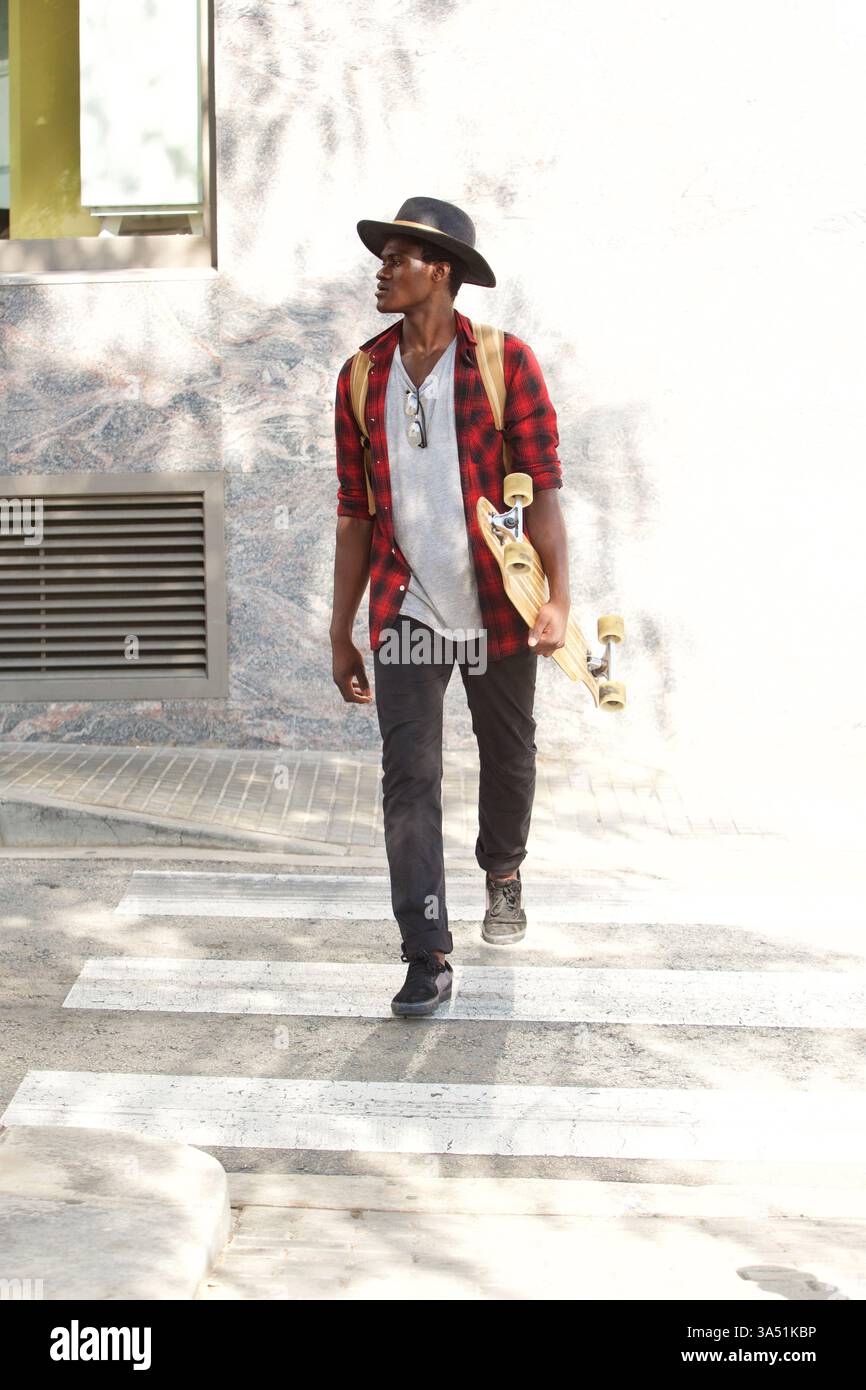 Stilvoller junger Mann, der draußen mit einem Skateboard spaziert, aufgenommen in einem zeitgenössischen Straßenbild. Perfekt für Lifestyle- und Modethemen mit lässigem Selbstbewusstsein. Stockfoto