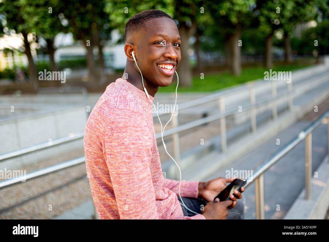 Seitenporträt eines jungen Schwarzen Mannes, der draußen Musik mit Ohrhörern hört. Er lächelt, während er sein Handy in einer lebhaften städtischen Umgebung ansieht. Stockfoto