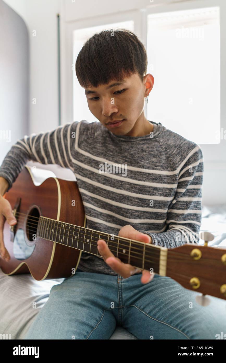 Asiatischer Mann spielt Gitarre, während er auf einem Bett in einem sonnendurchfluteten Schlafzimmer sitzt. Zwangloses Zuhause legt Wert auf Hobby und Entspannung. Ideal für Musik, Lernen und Bilder im Heimleben. Stockfoto