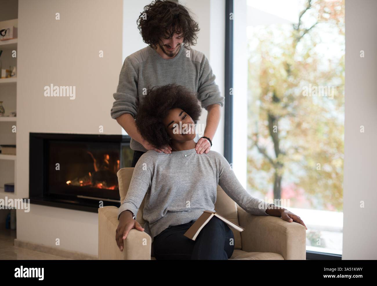 Warme, intime Szene eines jungen multiethnischen Paares, das an einem Herbsttag zu Hause an einem gemütlichen Kamin umarmt. Sie lesen zusammen ein Buch, lächeln und entspannen sich in einer komfortablen Inneneinrichtung. Dieses romantische Lifestyle-Bild unterstreicht die Wärme, Zweisamkeit und saisonale Gemütlichkeit. Stockfoto
