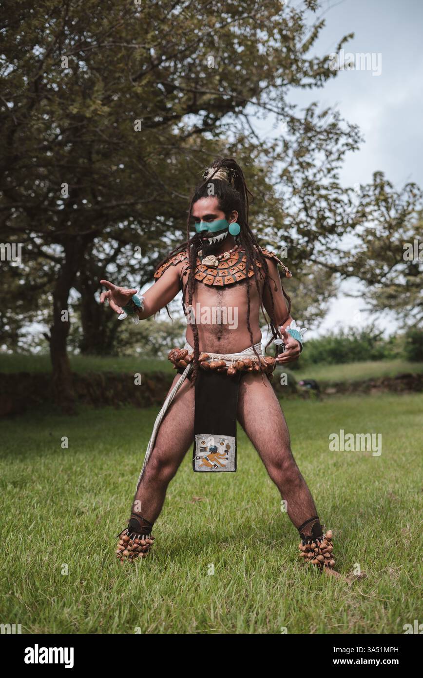 Hispanischer Mann mit Gesichts- und Körpermalereien in traditionellem Maya-Kriegerkostüm, der für den Kampf trainiert und wegblickt, während er auf Gras ... steht Stockfoto