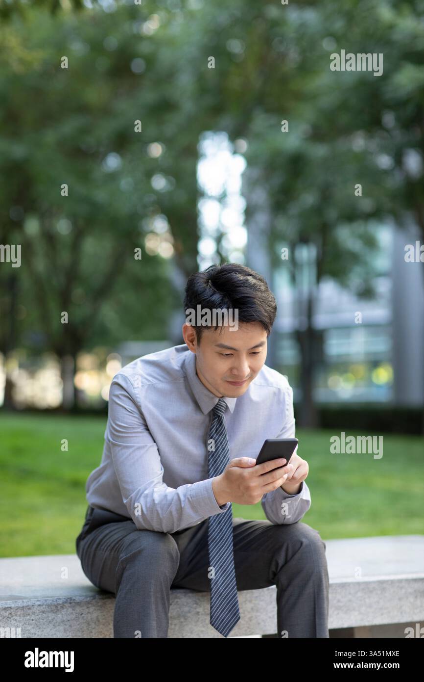Ein lächelnder chinesischer Geschäftsmann verwendet tagsüber ein Smartphone, während er auf einer Bank im Park sitzt. Dieser entspannte Moment unterstützt mobiles Business, Konnektivität und den Outdoor-Lebensstil. Hervorragend geeignet für Technik und urbane Lebensfunktionen. Stockfoto