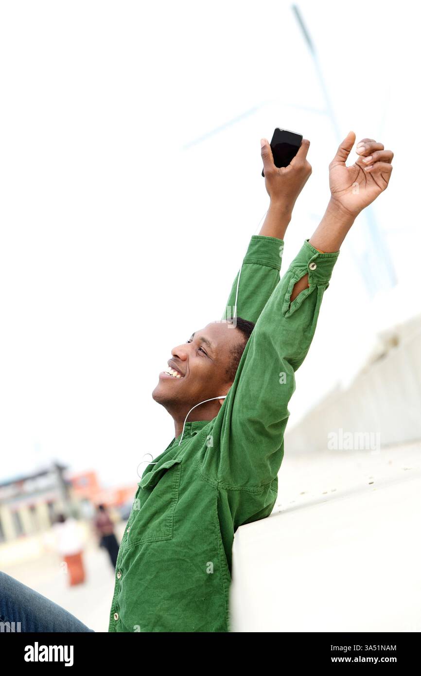 Porträt eines glücklichen jungen Mannes, der mit erhobenen Armen und einem Handy lächelt. Ein unbeschwertes, legeres Lifestyle-Image, das perfekt für mobile Technologien, Musik und Kommunikationsthemen geeignet ist. Geeignet für soziale, digitale und Lifestyle-Kampagnen. Stockfoto