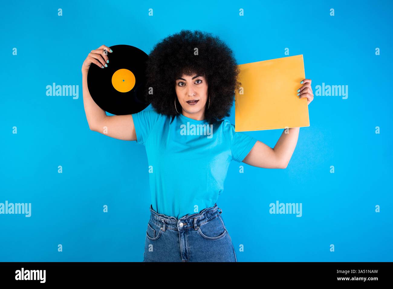 Lächelnde hispanische Frau mit einem Afro, der eine Retro-Schallplatte und ein Cover vor blauem Hintergrund hält. Dieses Vintage-Musikthema eignet sich hervorragend für Lifestyle- oder Musikkampagnen und Branding. Die Einstellung Clean Studio bietet Kopierraum für Text oder Grafiken. Stockfoto