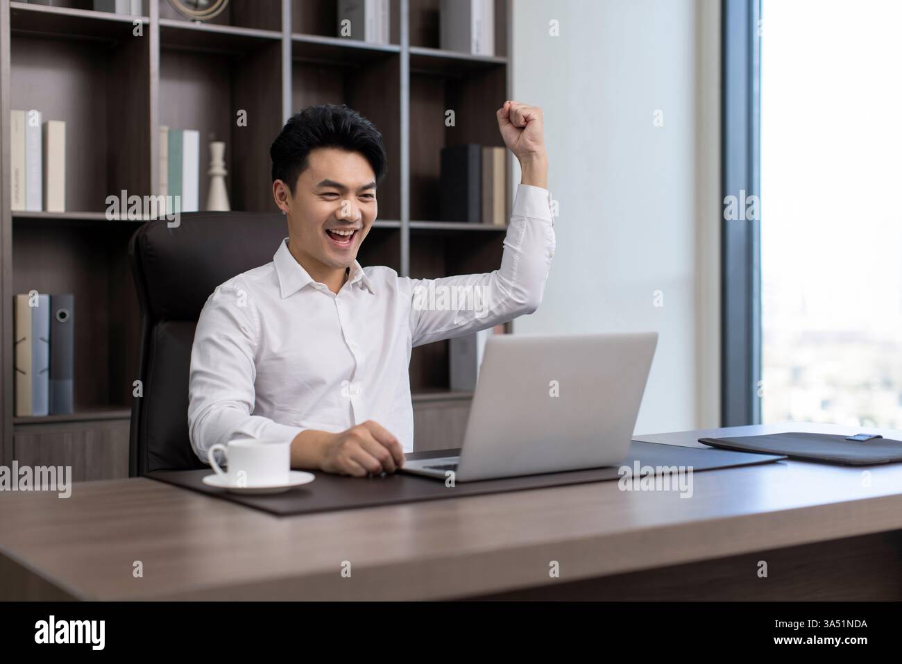 Der fröhliche chinesische Geschäftsmann feiert einen Erfolg, während er an einem Schreibtisch im Büro sitzt. Er hebt den Arm als Zeichen des Sieges neben seinem Laptop und vermittelt Vertrauen und Leistung. Dieses Image eignet sich ideal für Geschichten über Führung, Erfolg und Unternehmenswachstum. Stockfoto