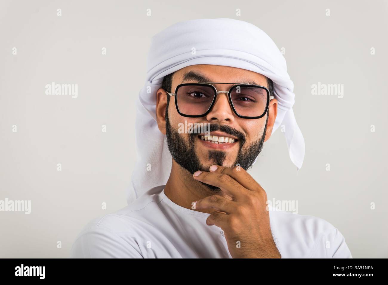 Studio-Porträt eines selbstbewussten Mannes aus dem Nahen Osten, der Kandura und Sonnenbrille trägt und eine durchdachte Hand am Kinn trägt. Geeignet für Geschäfts-, Reise- und Lifestyle-Kampagnen mit regionalem Stil. Weißer Hintergrund schafft sauberen Kopierraum für Überschriften. Stockfoto