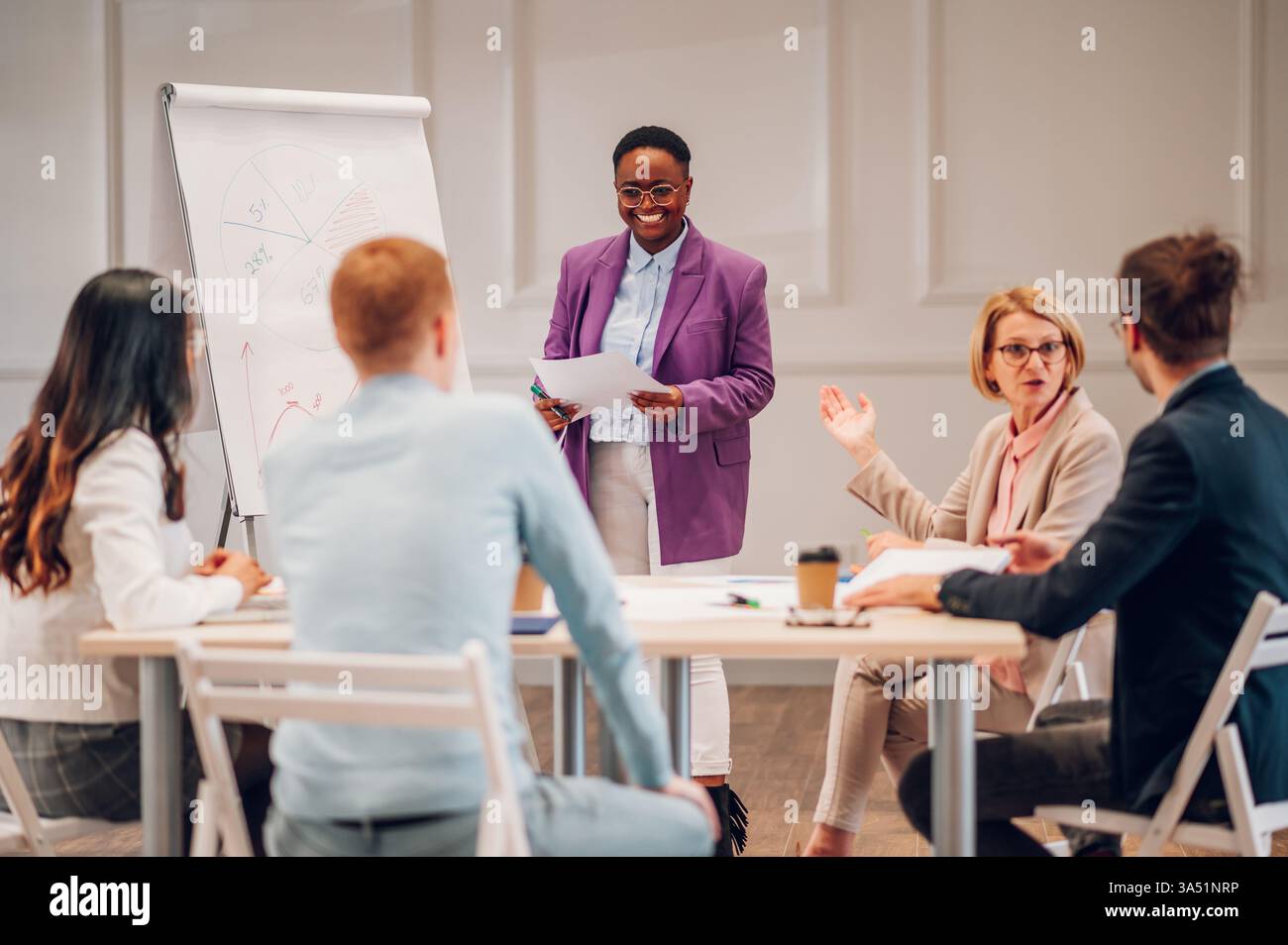 Schwarze Geschäftsfrau, die ihrem Team Marketingforschung in einem modernen Sitzungssaal vorstellt. Sie erläutert Vertriebsdaten mit Diagrammen und einem Whiteboard und erfasst Führungsqualitäten und Teamarbeit. Geeignet für Unternehmenskommunikation, weibliche Führungskräfte und Funktionen für Geschäftsstrategien. Stockfoto