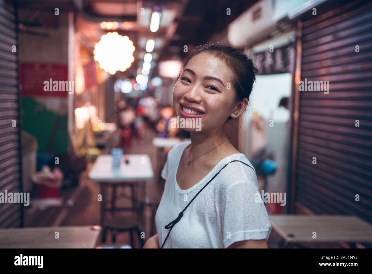 Fröhliche junge asiatische Frau in Freizeitkleidung lächelt nachts neben einem beleuchteten Straßencafé. Diese urbane Lifestyle-Aufnahme passt zu Street-Style, Nachtleben und zeitgenössischem Lifestyle. Stockfoto