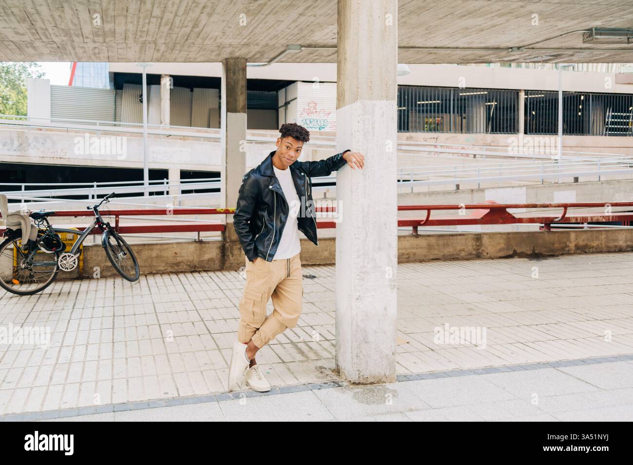 Schwarzer Mann, der sich an einen Pfosten lehnt, mit der Hand in der Tasche, draußen bei Tageslicht. Lässiges Porträt im Street-Stil mit Lederjacke. Ideal für Lifestyle, Outdoor-Mode und urbane Wohnmotive. Stockfoto