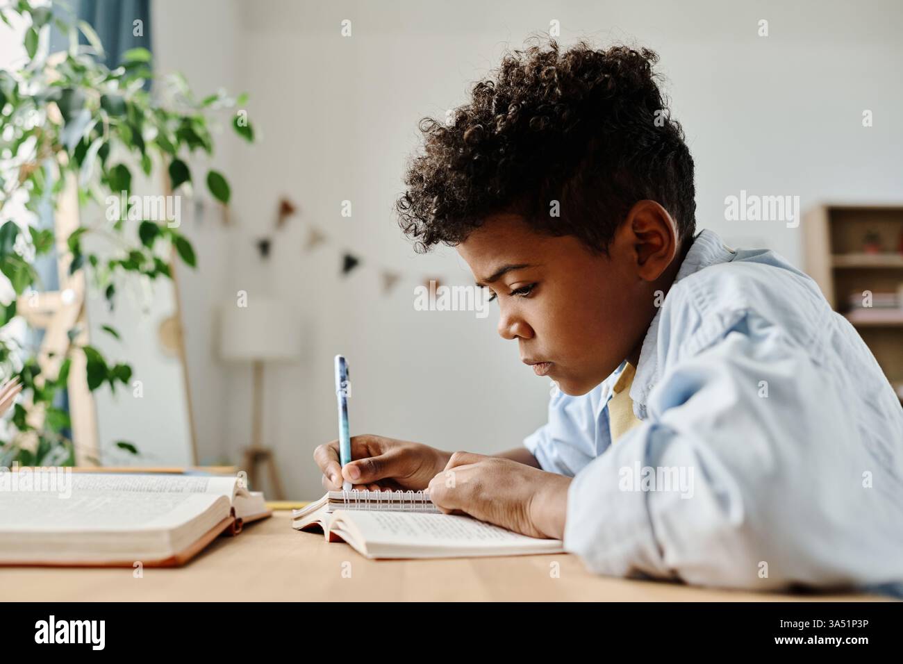 Afrikanischer Grundschüler, der Hausaufgaben zu Hause macht, an einem Tisch mit Buch und Notizbuch sitzt. Dieser konzentrierte Lernmoment spiegelt Bildung und Unterstützung durch die Familie wider. Stockfoto