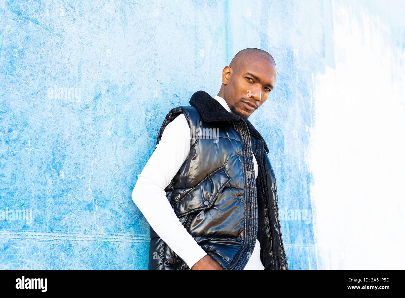 Ein entspannter Afro-Latino-Mann lehnt sich an eine Betonmauer in einer städtischen Tagesumgebung. Der Street-Style-Look und die selbstbewusste Pose passen zu Lifestyle-, Mode- und urbanen Lifestyle-Themen. Ideal für Marketinginhalte, die sich auf verschiedene männliche Profis oder das Stadtleben konzentrieren. Stockfoto