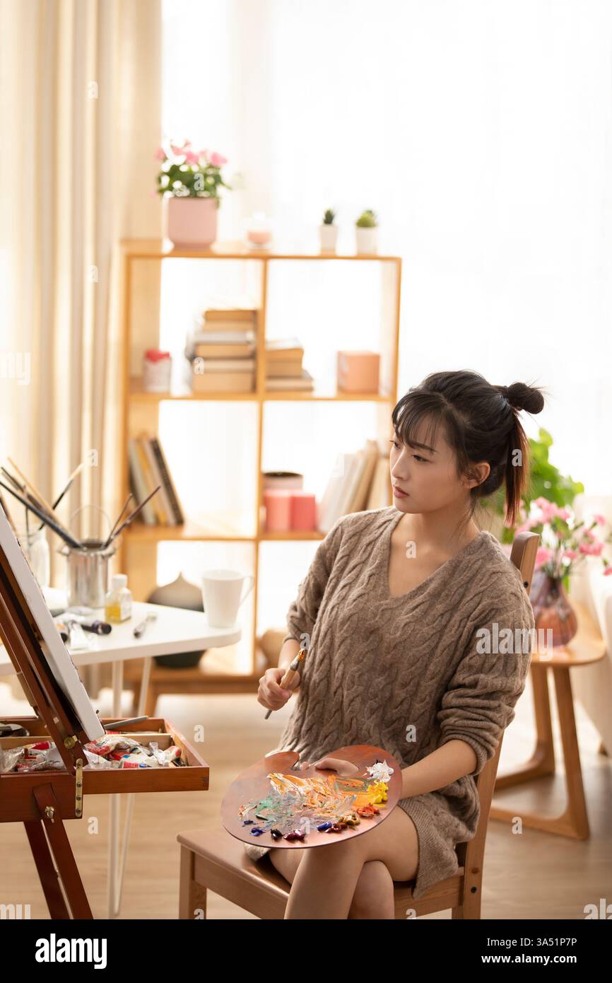 Chinesin malt auf Leinwand, während sie auf einem Stuhl in einem Heimstudio sitzt. Ihre Staffelei und Malerei heben Kreativität, Kunst und Handwerk sowie das häusliche Leben hervor. Perfekt für Darstellungen zu Künstlern, Hobbys und Kunstprojekten. Stockfoto