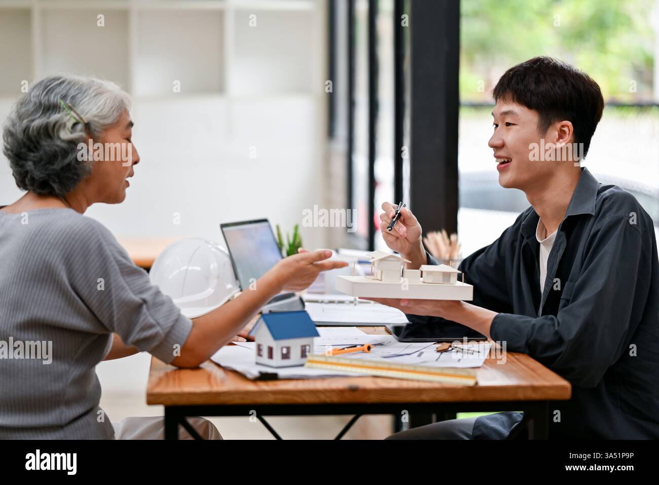 Ältere asiatische Frau, die sich mit einem asiatischen Ingenieur trifft, der das Modellhaus hält und bespricht, während sie am Tisch im Büro sitzt Stockfoto