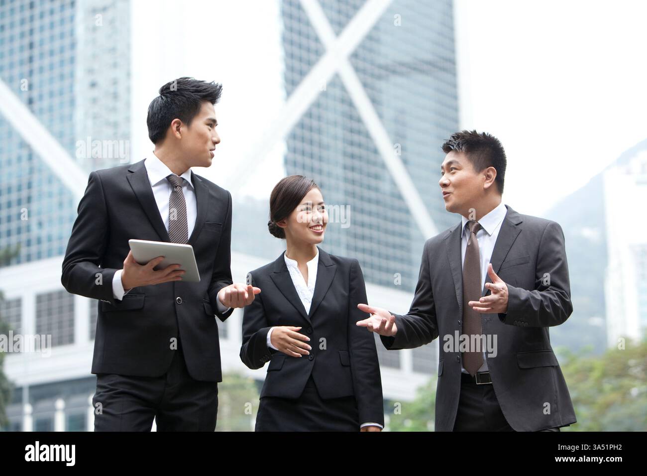 Fröhliche chinesische Geschäftsleute unterhalten sich in formeller Kleidung, während sie eine Stadtstraße entlang laufen. Der Moment vermittelt Networking, Teamwork und positive Geschäftsdynamik in einem städtischen Umfeld. Nützlich für Geschichten über Partnerschaft und modernen Handel. Stockfoto