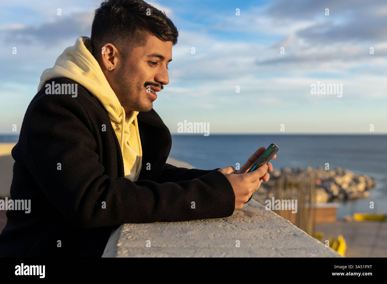 Lifestyle-Aufnahme eines glücklichen hispanischen Mannes in Oberbekleidung, der lächelt, während er auf seinem Smartphone am Meer in den sozialen Medien blättert. Die Seitenansicht zeigt ihn, wie er sich auf einen Damm mit dem Wasser im Hintergrund lehnt, was eine entspannte urbane Küstenatmosphäre erzeugt. Ideal für Lifestyle- und Mobiltechnologie-Geschichten mit Online-Nachrichten und lässigen Momenten am Meer in der Abenddämmerung. Stockfoto