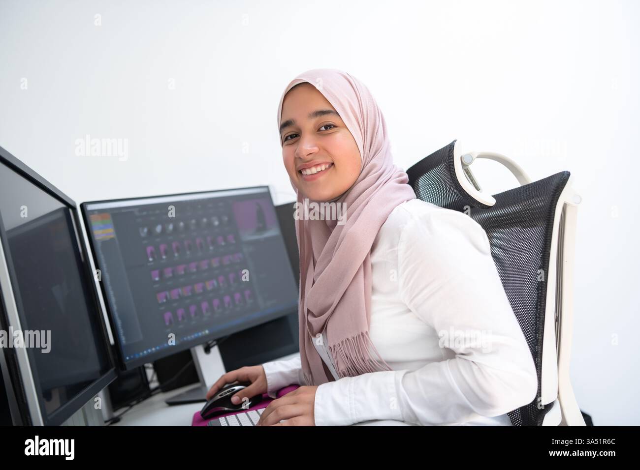 Weibliche arabische Kreativprofi, die in einem Heimbüro mit zwei Monitoren arbeitet und einen Hijab trägt. Eine moderne Szene, in der Unternehmergeist, Technologie und fokussierte Produktivität in einem modernen Interieur hervorgehoben werden. Stockfoto